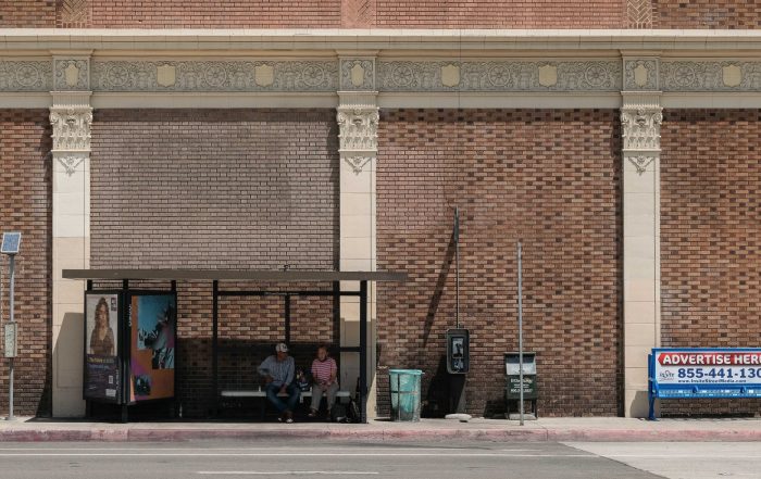 A man and a woman sit beneath the shade of a bus shelter on a sunny day.