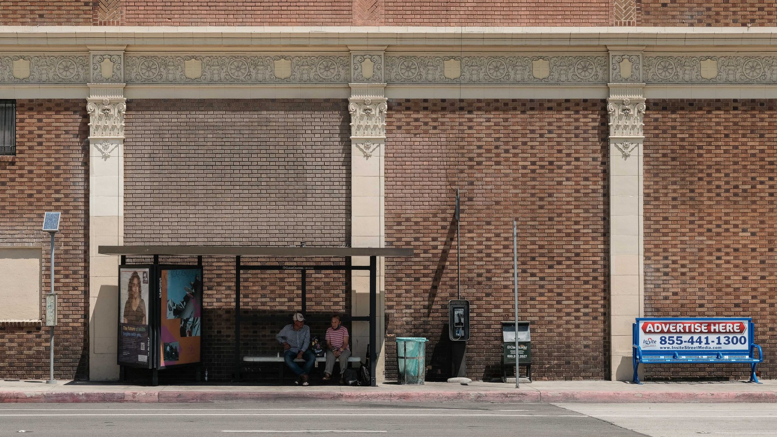 A man and a woman sit beneath the shade of a bus shelter on a sunny day.