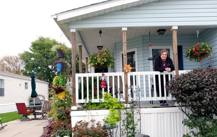 An older woman smiles from the porch of a light blue manufactured home, surrounded by hanging flower baskets and a vibrant garden. The porch is decorated with bird feeders and plants. Patio furniture and another home are visible in the background.