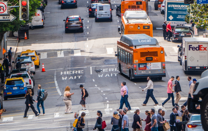 A busy street in downtown los angeles shows pedestrians crossing the street, buses on the road, along with cars, taxis and delivery vehicles.