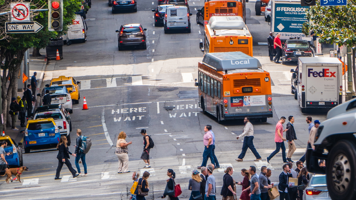 A busy street in downtown los angeles shows pedestrians crossing the street, buses on the road, along with cars, taxis and delivery vehicles.